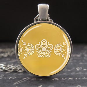 May include: A silver-toned pendant necklace featuring a round, mustard-yellow pendant. The pendant has a white floral design in the center and along the edges. The necklace chain is also silver-toned.