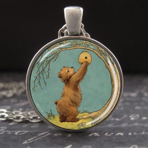 May include: A silver-toned pendant necklace featuring a round charm with a whimsical illustration. The image depicts a brown bear reaching for a yellow and brown beehive hanging from a tree branch, set against a teal background.
