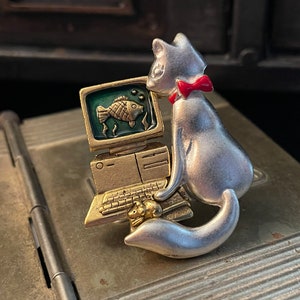 May include: A silver cat figurine wearing a red bow tie, looking at a gold computer with a green screen and a fish on it.