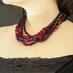 May include: A multi-strand necklace made of yarn in shades of red, purple, and blue. The necklace is woven in a braided pattern and is worn by a person.