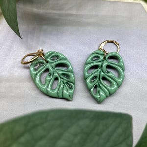 May include: A pair of pale green leaf-shaped earrings with gold-coloured hoops. The earrings are a detailed replica of a Monstera leaf, with intricate cut-out patterns. The earrings are set against a neutral background with green foliage.