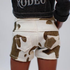 May include: Doll-sized white denim shorts with brown cow print spots and two back pockets. The shorts are paired with a black denim jacket with the word "RODEO" on the back.