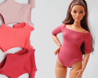 May include: A doll wearing a pink off-the-shoulder bodysuit, with three additional bodysuits in shades of pink. The doll has long brown hair and is posed against a white background. The bodysuits are made of a soft fabric.