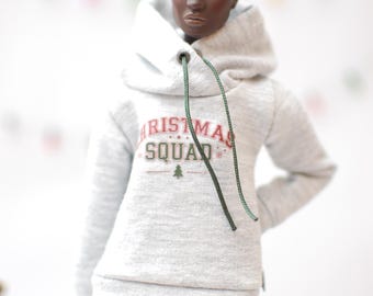 May include: A doll wearing a light gray hoodie with the text "CHRISTMAS SQUAD" in red and green. The doll is also wearing patterned leggings with red, white, and green snowflake and Christmas tree designs. The hoodie has a drawstring.