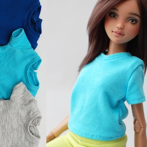 May include: Three plain t-shirts in blue, turquoise, and gray stacked on top of each other. A doll wearing a turquoise t-shirt and yellow pants is in the background.