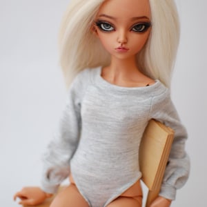 May include: A doll with long, light blonde hair and detailed facial features, wearing a gray bodysuit with long sleeves. The doll is seated on a small wooden chair, with a neutral background.