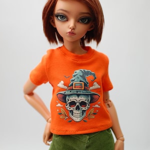 May include: A doll wearing an orange t-shirt with a skull and witch hat graphic. The doll has brown hair and is wearing green corduroy pants.