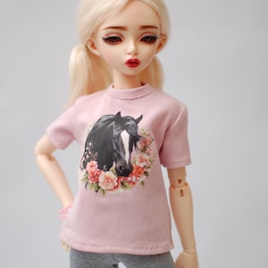 May include: A pink t-shirt with a black horse and floral design. The doll is wearing grey leggings.