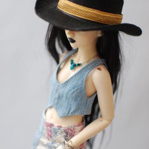 May include: A doll wearing a black hat with a gold band, a blue denim vest, and a floral print skirt. The doll has black lipstick, a turquoise necklace, and silver bracelets. The hat's brim casts a shadow over the doll's face.