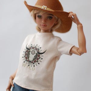 May include: A doll wearing a brown cowboy hat and a cream-colored t-shirt with a bull skull graphic. The doll is also wearing blue jeans. The shirt has the word "WILD" printed below the graphic.