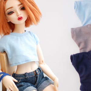 May include: A doll with orange hair and red lipstick wearing a light blue crop top and denim shorts. Three additional t-shirts in light blue, taupe, and navy blue are stacked in the background. The doll is seated on a small wooden chair.