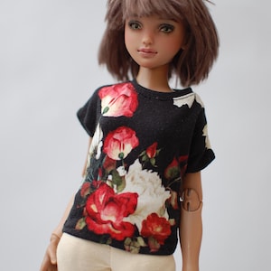 May include: A doll wearing a black t-shirt with a vibrant floral pattern of red roses and white flowers. The doll also wears cream-colored pants. The doll has brown hair and is set against a plain white background.