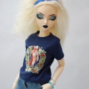 May include: A doll with blonde hair wearing a blue headband, a navy blue t-shirt with a horse graphic, and blue jeans.