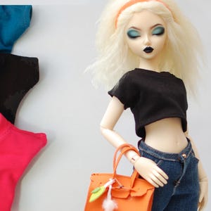 May include: A doll wearing a black crop top, blue jeans, and carrying an orange handbag. The doll has blonde hair and is wearing a headband. Several colorful t-shirts are visible in the background. The doll's makeup includes blue eyeshadow and black lipstick.