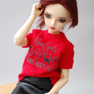 May include: A doll with red hair wearing a red t-shirt with the text "Bad Girls Have More Fun" and a black skirt. The doll is sitting on a small wooden chair.