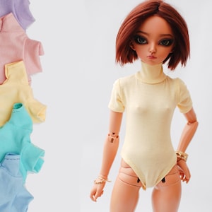 May include: A doll wearing a yellow turtleneck bodysuit with short sleeves. The doll has long, straight, auburn hair and brown eyes. The doll is wearing a gold bracelet on its left wrist.