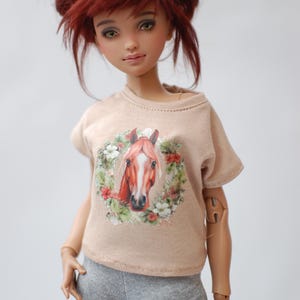 May include: A doll with red hair in two buns wearing a beige t-shirt with a horse print and grey leggings.