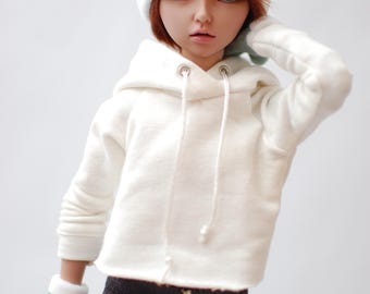 May include: A doll wearing a white hoodie, teal and white beanie, and matching gloves. The doll has red hair and is wearing dark leggings with a maroon pattern. The doll is posed with one hand on its head.