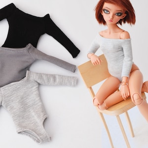 May include: Two gray and one black off-the-shoulder long-sleeve tops for dolls. The tops are made of a soft, stretchy fabric. The doll is wearing a gray top and sitting on a wooden chair.