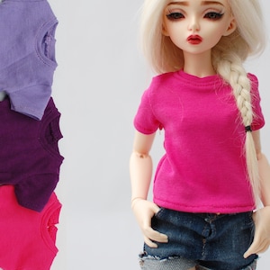 May include: A doll wearing a pink short-sleeved t-shirt and denim shorts. Three t-shirts in purple, pink, and lavender are stacked behind the doll.