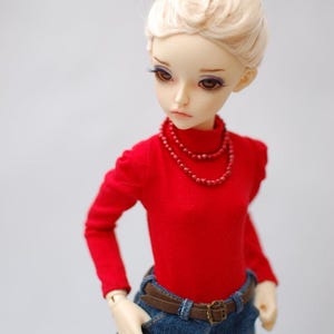 May include: A doll with blonde hair styled in a bun, wearing a red long-sleeved top, blue jeans, and a brown belt. The doll has a red beaded necklace. The doll's skin is pale, and it has brown eyes.