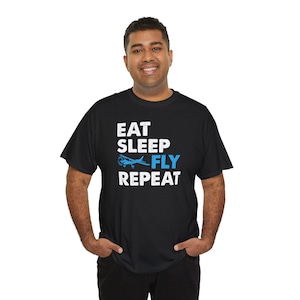 Op de afbeelding: Zwart T-shirt met witte tekst die "Eat Sleep Fly Repeat" zegt en een blauwe vliegtuigafbeelding.