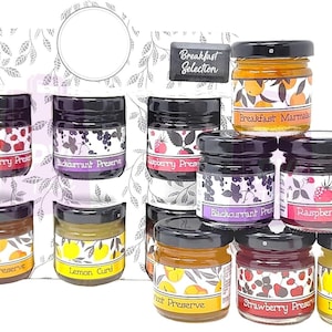 May include: A selection of eight small glass jars filled with various preserves and marmalades. The jars are arranged in two rows of four, with labels that read "Strawberry Preserve", "Blackcurrant Preserve", "Blueberry Preserve", "Apricot Preserve", "Lemon Curd", "Blackcurrant Preserve", "Raspberry Preserve", and "Strawberry Preserve". The jars are labelled "Breakfast Selection".