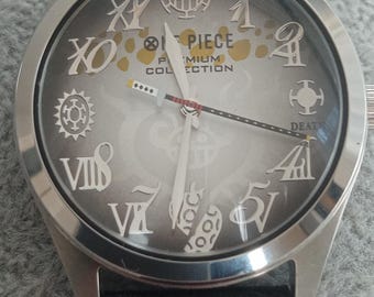 Könnte beinhalten: Eine silberne Armbanduhr mit schwarzem Lederarmband. Das graue Zifferblatt der Uhr zeigt silberne römische Ziffern und Symbole. Die Worte "ONE PIECE PREMIUM COLLECTION" sind auf dem Zifferblatt aufgedruckt.