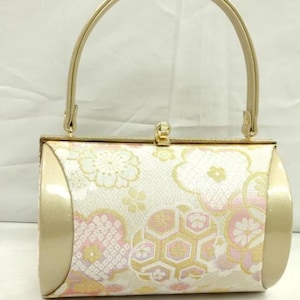May include: A cylindrical handbag with a gold handle and accents. The body of the bag features a floral pattern in white, pink, and gold. The bag has a clasp closure.