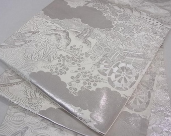 May include: A white fabric with a silver metallic sheen. The fabric has a repeating pattern of stylized flowers, leaves, and a cart with a wheel.