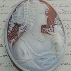 Op de afbeelding: Ovale camee met een vrouw in profiel met krullend haar, een parelketting en een vogel op haar hand. De camee is wit met een bruine achtergrond en is gesigneerd "Agretti".
