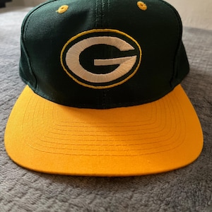 Puede incluir: Gorra de béisbol verde y amarilla con una "G" bordada en blanco en la parte delantera.