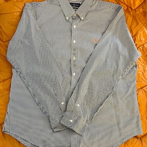 May include: A long-sleeved button-down shirt with a small, checkered pattern in navy blue and white. The shirt has a button-down collar, a chest pocket, and a small orange logo on the chest. The shirt is laid flat on an orange surface.