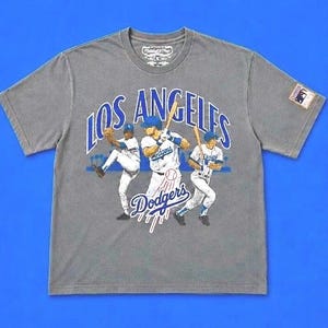May include: A grey short-sleeve t-shirt featuring a graphic of three baseball players in white uniforms. The text LOS ANGELES is printed in blue above the players, with Dodgers written in blue script below. A small MLB patch is on the left sleeve.