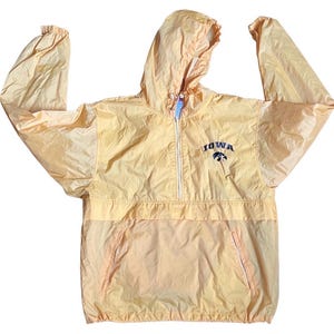 Könnte beinhalten: Eine hellgelbe, langärmelige Windjacke mit Kapuze, einem halben Reißverschluss vorne und elastischen Bündchen. Auf der Brust befindet sich der gestickte Schriftzug "IOWA" in Dunkelblau über einem kleinen, dunkelblau-gelben Falken-Logo.