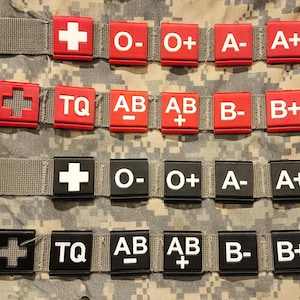 May include: A collection of red and black medical identification patches with white lettering and symbols, including blood types and a cross symbol. The patches are attached to a camouflage background.