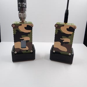 May include: Two electronic radio devices with camouflage patterned casings, each mounted in a black base. The left device features a tall black antenna labeled ALFA 915, while the right device has a thinner black antenna. Both units have silver screws.
