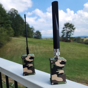 May include: Two small, rectangular devices with camouflage patterns. One has a thin, black antenna, the other a thicker, black antenna. Both are on a white railing, with a green landscape and blue sky in the background.