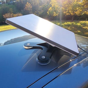 May include: A rectangular, white satellite dish mounted on a black bracket, attached to a blue car roof. The dish is angled upwards, with a cable running from it. The background shows trees and a grassy area.