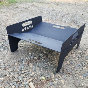 May include: Black metal fire pit with a rectangular shape and four legs. The fire pit has cutouts on the sides and a raised lip around the perimeter.