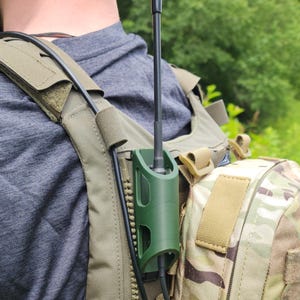 May include: A dark olive green plastic radio antenna holder mounted on a tan tactical vest. The holder secures a thin black antenna that extends vertically. A black cable runs along the side of the holder, which is attached to a camouflage pouch.