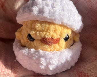 May include: A crocheted chick toy in a white eggshell. The chick is yellow with black eyes and a red beak. The eggshell is made of white yarn.