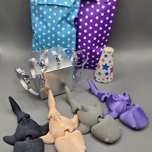 May include: A set of three 3D printed shark toys in purple, grey, and beige. The toys are posed in a playful manner, with their mouths open and their fins spread out. The toys are surrounded by gift wrapping supplies, including a silver ribbon, a blue and white polka dot gift bag, a purple polka dot gift bag, and a colourful party hat.