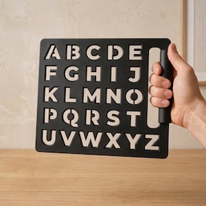 May include: Black alphabet stencil with a handle. The stencil features the letters A through Z in a sans-serif font. The letters are cut out, allowing for tracing or painting. The stencil is held by a person.