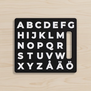 May include: A black alphabet board with white uppercase letters. The letters are arranged in rows and columns, with a handle cutout on the right side. The board includes the letters Å, Ä, and Ö.