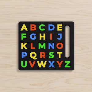 May include: A black square board featuring the English alphabet in raised, colorful letters arranged in a grid. The letters A through Z are displayed in yellow, orange, green, and blue. A vertical rectangular cutout is located on the right side.