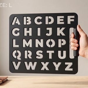 May include: A black rectangular stencil featuring the alphabet from A to Z in a bold, sans-serif font. The stencil includes a vertical handle on the right side and the text SIZE: L printed in the top left corner. The letters are cutouts.