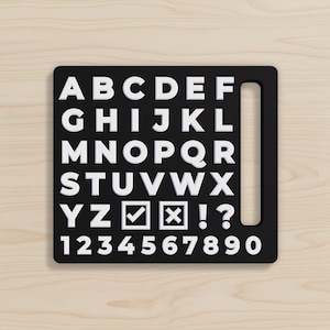 May include: A black rectangular stencil featuring white raised letters from A to Z, numbers 0 through 9, a checkmark symbol, an X symbol, an exclamation point, and a question mark. The stencil includes a vertical handle slot on the right side.