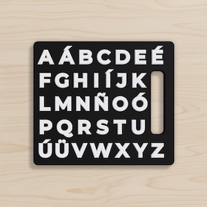May include: A black wooden alphabet puzzle with white letters. The letters are arranged in rows and columns, with a cutout on the right side. The letters include accented characters. The puzzle is on a light wood surface.