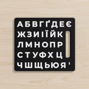 Puede incluir: Una tabla rectangular negra con letras cirílicas blancas dispuestas en filas. La tabla tiene un recorte en forma de asa en el lado derecho. Las letras son en negrita, sin serifa.